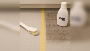 ユニットバスの床を中央のテープで区切り、左は清掃後で明るく、右は黒ずみが残る様子。手前にブラシ、右上に「中性」洗剤ボトルを収めた構図