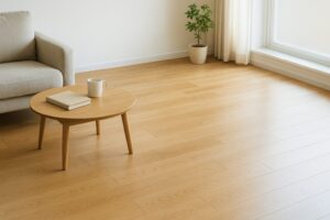 日差しの入るリビングをやや俯瞰で。手前から奥へオーク系フローリングの木目が伸び、左手前に低めの布ソファ、中央に丸い木製ローテーブル。