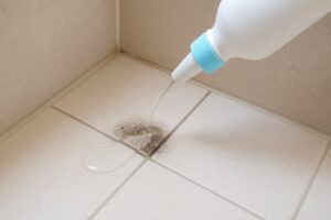 風呂の床の隅に液体の漂白剤が透明な状態で撒かれている場面。床はベージュ系のタイルで、窓からの自然光が差し込み、淡い陰影が見える清潔感のある実写構図。