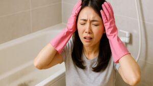 ピンクのゴム手袋をはめた若い日本人女性が浴室で頭を抱える「オーマイガー」ポーズ。背後に浴室タイルと浴槽が映り、風呂の黒ずみが落ちずに困っている様子が伝わる。