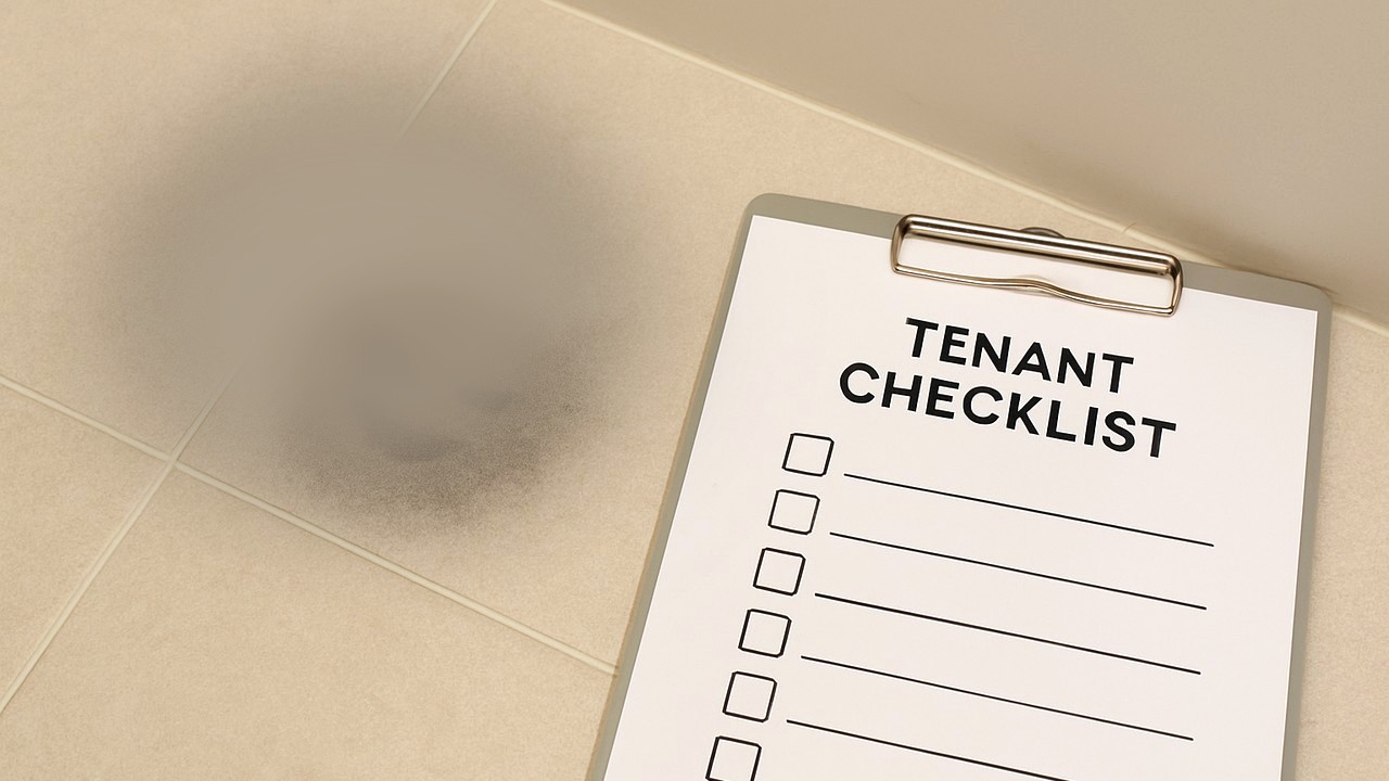 ベージュ寄りに明るい浴室床タイルの一角に、広く薄く黒ずみが残っており、その右側に「TENANT CHECKLIST」と書かれた退去チェックリストのクリップボードが置かれている実写写真。