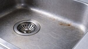 細かい傷と点状のサビが浮いたステンレス製キッチンシンクの底面と排水口がアップで写っており、くすんで輝きを失った状態がわかる写真